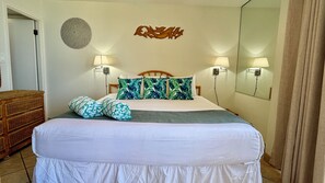 Un lit bien fait, avec une couette blanche, deux coussins verts à motifs et un plaid gris. Deux lampes murales sont fixées de chaque côté du lit.