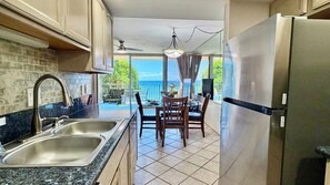 A kitchen with a stainless steel refrigerator, a double sink, and a dining area with a table and chairs. - Welcome to our beachfront Maui 'Ohana! - Steps to the beach! (Lahaina)