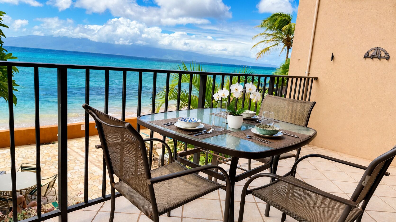 A balcony with a table set for dining, overlooking a beach with clear blue water and a palm tree.