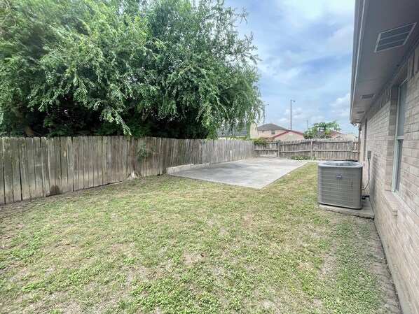A backyard with a wooden fence, a grassy area, and a large air conditioning unit.
