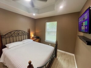 Een slaapkamer met een bed, nachtkastje, lamp, raam met zonwering en een aan de muur bevestigde televisie.