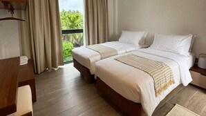 Ein Hotelzimmer mit zwei Betten, einem Schreibtisch, einem Stuhl, einem Deckenventilator und Blick auf Grünflächen.