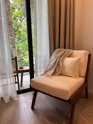 Une pièce dotée d’un plancher en bois, d’un canapé beige agrémenté d’un plaid à motifs et offrant une vue sur l’extérieur à travers une fenêtre ornée de rideaux légers.