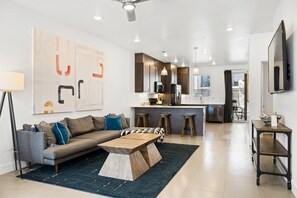 A modern living room with a sofa, coffee table, and bar stools. There is a kitchen area with a refrigerator, microwave, and sink.