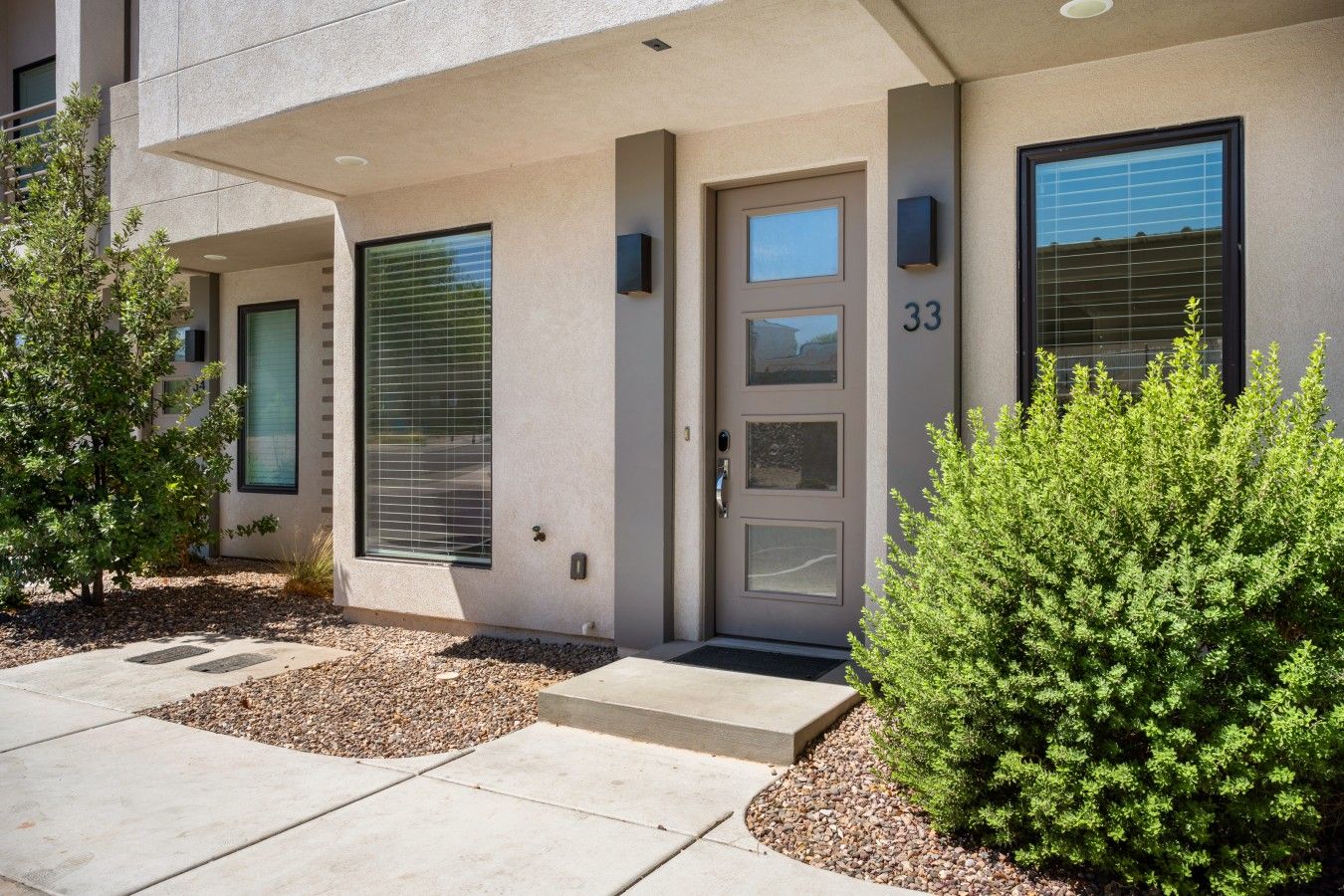 A modern building with a grey facade, a numbered door, and a small garden area with shrubs.