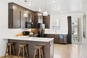 A modern kitchen with dark wood cabinets, stainless steel appliances, a white island with stools, pendant lighting, and a view of the outdoors. - Poolside Family Retreat, Sleeps 14 (St. George)