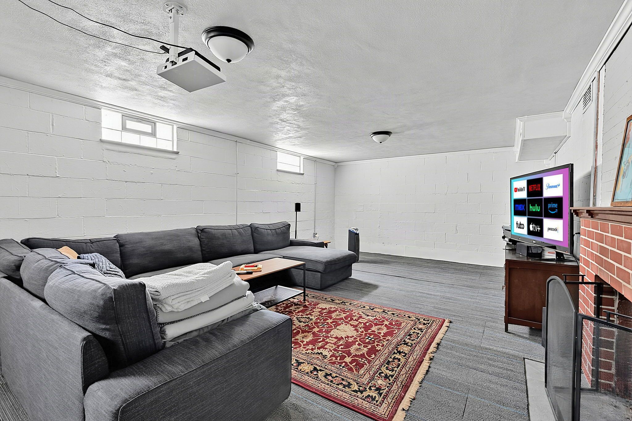 A modern living room with a gray sofa, a red patterned rug, a flat-screen TV displaying streaming services, and a ceiling-mounted projector.