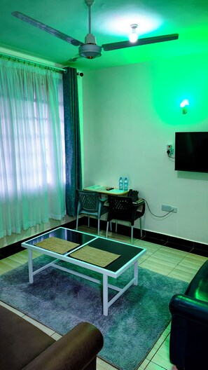 Una sala de estar con ventilador de techo, televisor de pantalla plana montado en la pared, una mesa de centro con papeles, una silla y un sofá.