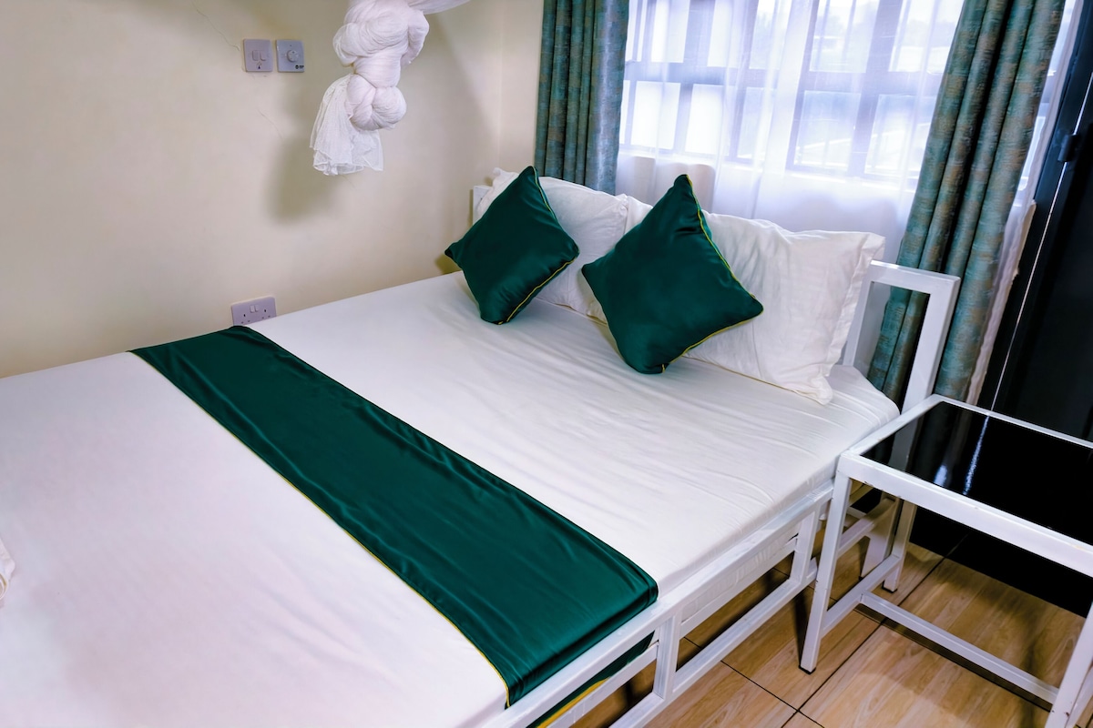 A neatly made bed with white bedding and a green throw, a white chair, a wall socket, and a window with curtains.