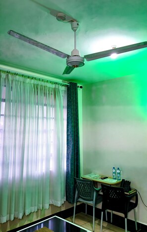 A room with a ceiling fan, a table with chairs, a water bottle, and a phone. - Aljona Apartments Hotel (Taveta)