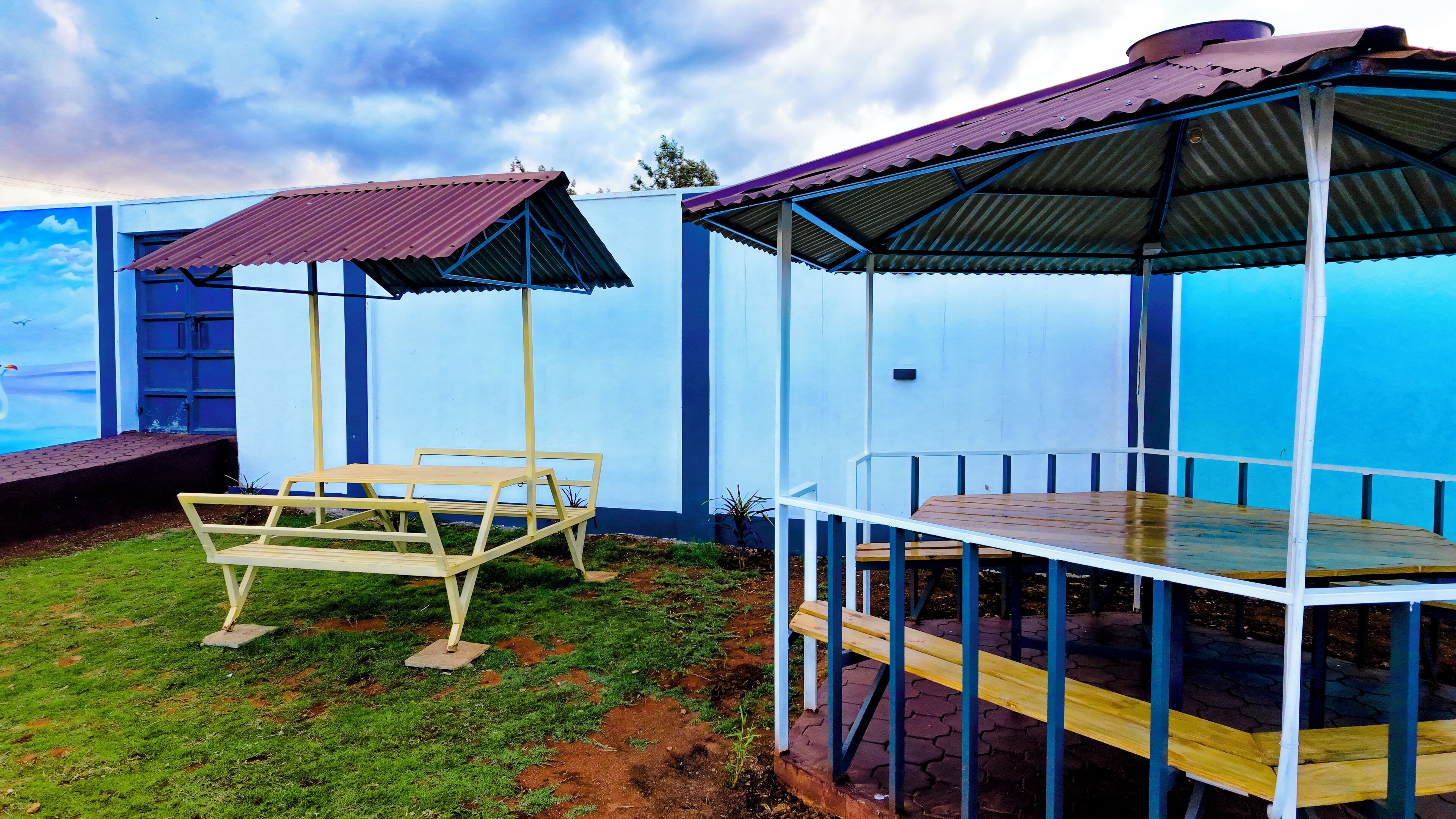 Un espacio de estar al aire libre cubierto, con una mesa, sillas, un pequeño jardín y un edificio de fachada azul.