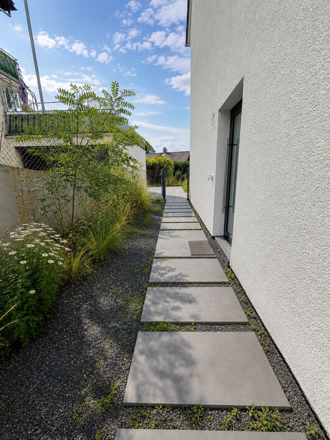 Ein schmaler Weg führt zu einem Gebäude mit einer strukturierten Außenwand und einer Tür.