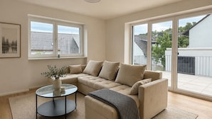 A modern living room with a beige sectional sofa, a glass coffee table, and a large window offering a view of a residential area. - The Golden Sunset Villa Wiesbaden (Schlangenbad)