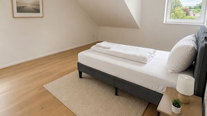 A bedroom with a bed, a bedside table with a lamp, a window with a view, and a framed picture on the wall. - The Golden Sunset Villa Wiesbaden (Schlangenbad)