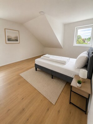 A bedroom with a bed, a bedside table with a lamp, a window with a view, and a framed picture on the wall. - The Golden Sunset Villa Wiesbaden (Schlangenbad)