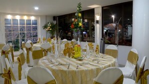 Un salón de banquetes con mesas redondas, sillas blancas y doradas, y centros de mesa florales.