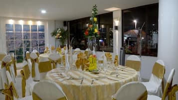 Un salón de banquetes con mesas redondas, sillas blancas y doradas, y centros de mesa florales.