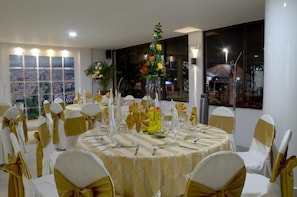 Un salón de banquetes con mesas redondas, sillas blancas y doradas, y centros de mesa florales.