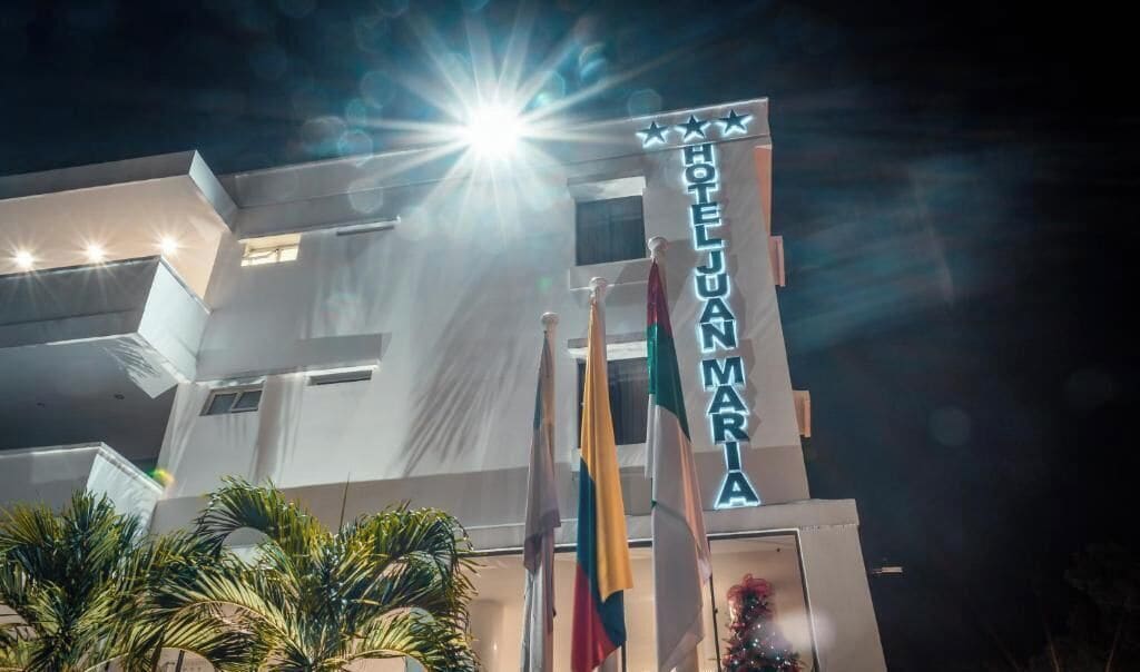A hotel named 'HOTEL JUAN MARIA' with a modern facade, multiple flags, and a bright light source in the background.