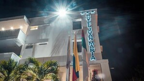 Un hotel llamado 'HOTEL JUAN MARIA' con una fachada moderna, varias banderas y una fuente de luz brillante al fondo.