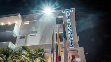 Un hotel llamado 'HOTEL JUAN MARIA' con una fachada moderna, varias banderas y una fuente de luz brillante al fondo.