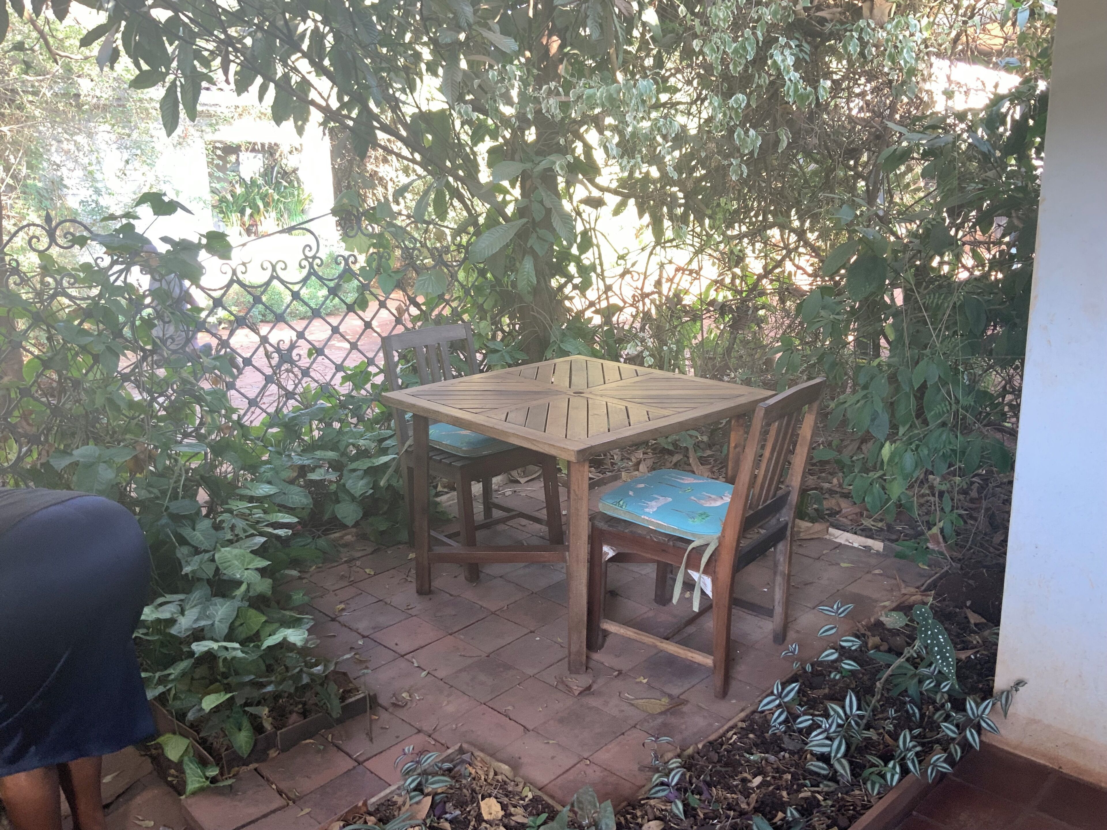 A wooden table and chairs set outdoors with a tiled floor, surrounded by greenery and a metal fence.