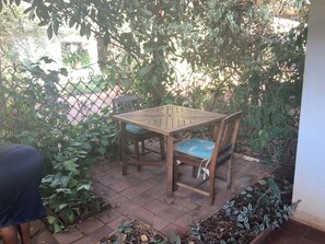 A wooden table and chairs set outdoors with a tiled floor, surrounded by greenery and a metal fence. - Adorable 2-bedroom bungalow in Nairobi perfect for relaxing stays (Nairobi)