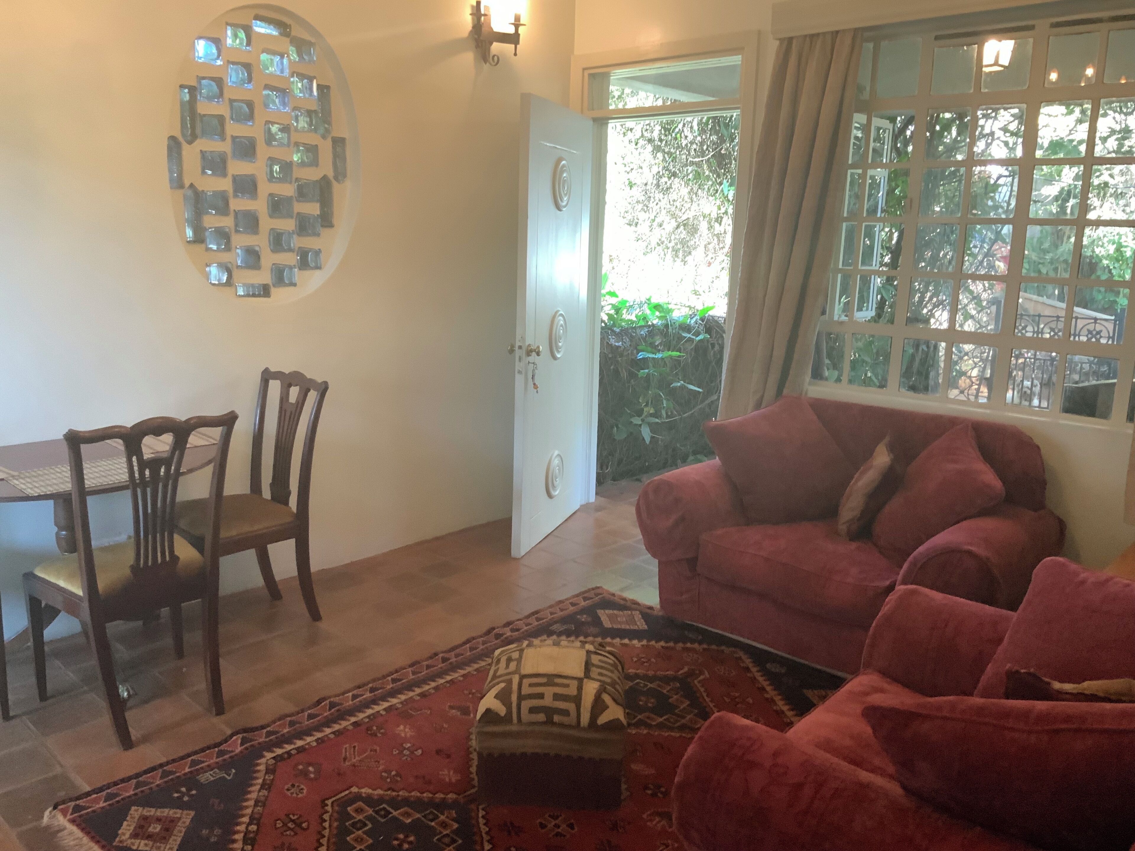 A room with a dining table, two chairs, a red sofa, a patterned rug, and a window with a view of greenery.