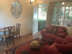 A room with a dining table, two chairs, a red sofa, a patterned rug, and a window with a view of greenery.