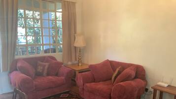 A living room with two red sofas, a small table with a lamp, a patterned rug, and a window with curtains.