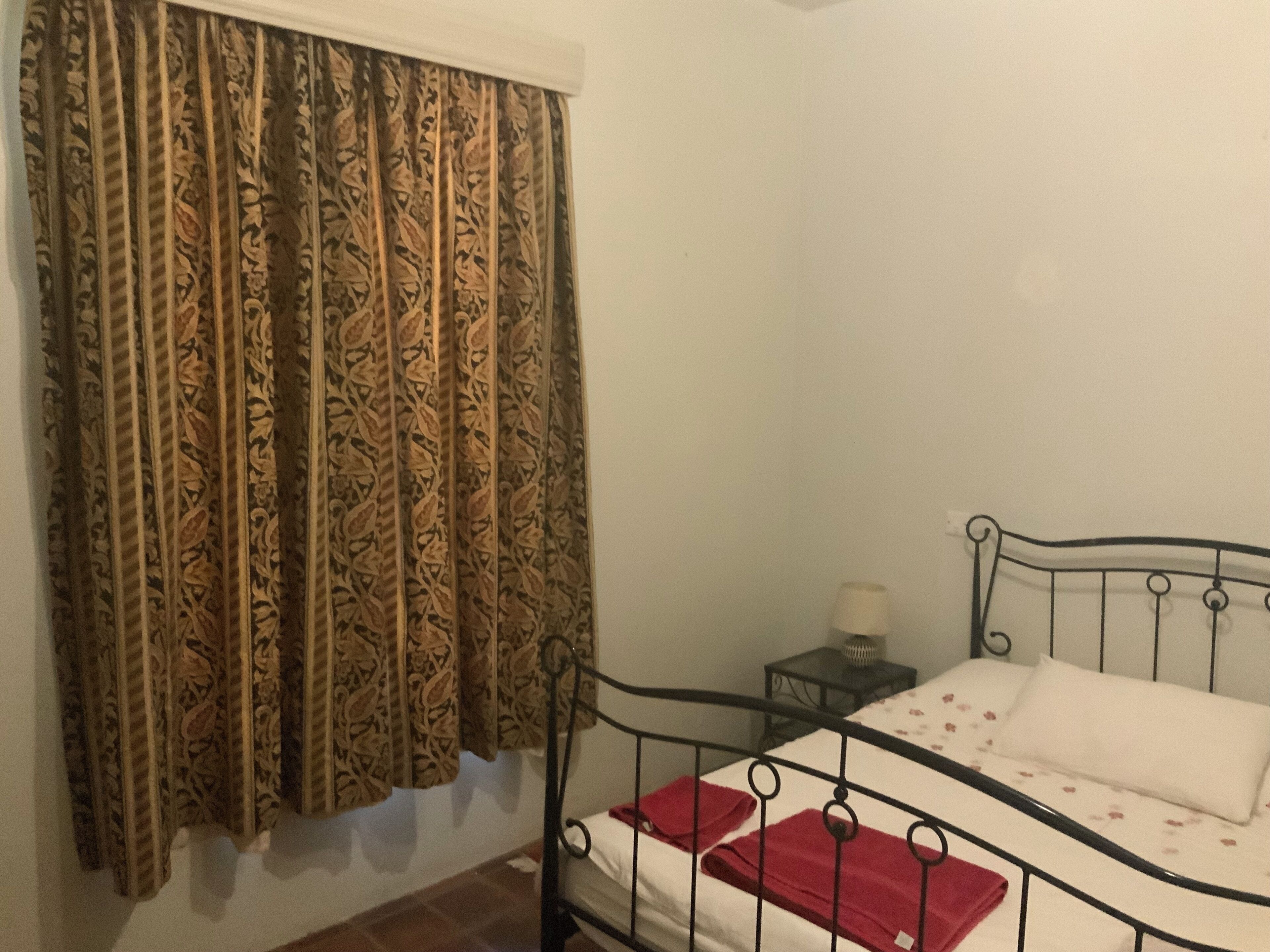 A single bed with a metal frame, a bedside table with a lamp, and a patterned curtain.