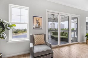 Una habitación con un sillón gris, un cuadro, vistas a un balcón con mobiliario exterior y una ventana con vistas al paisaje.