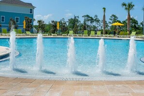 En swimmingpool med flera vattenstrålar, omgiven av gröna solstolar och gula parasoller, med en byggnad i bakgrunden.