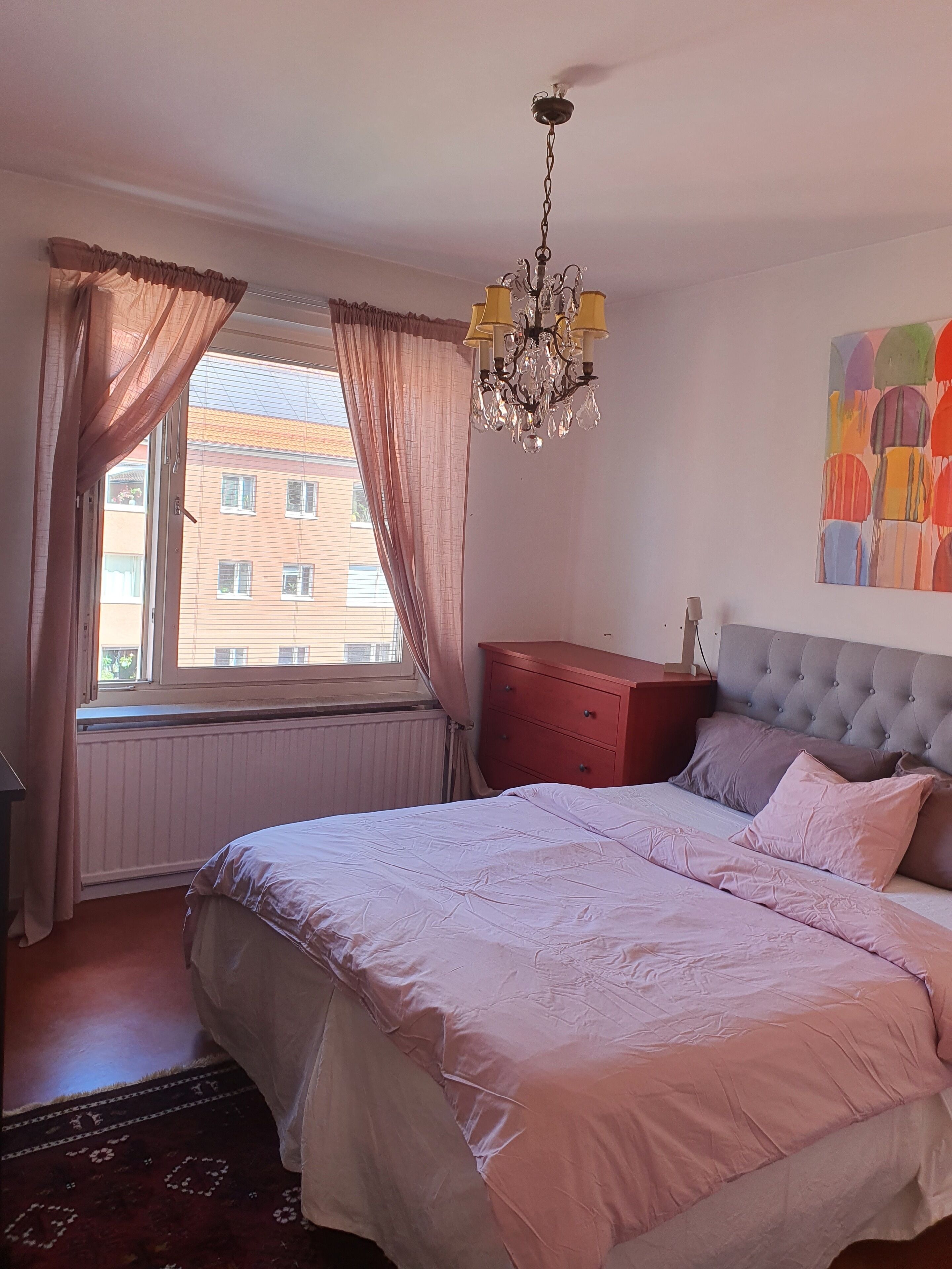 Ein Schlafzimmer mit Bett, Kommode, Heizkörper, einem Fenster mit Vorhängen und einer Chandelier.