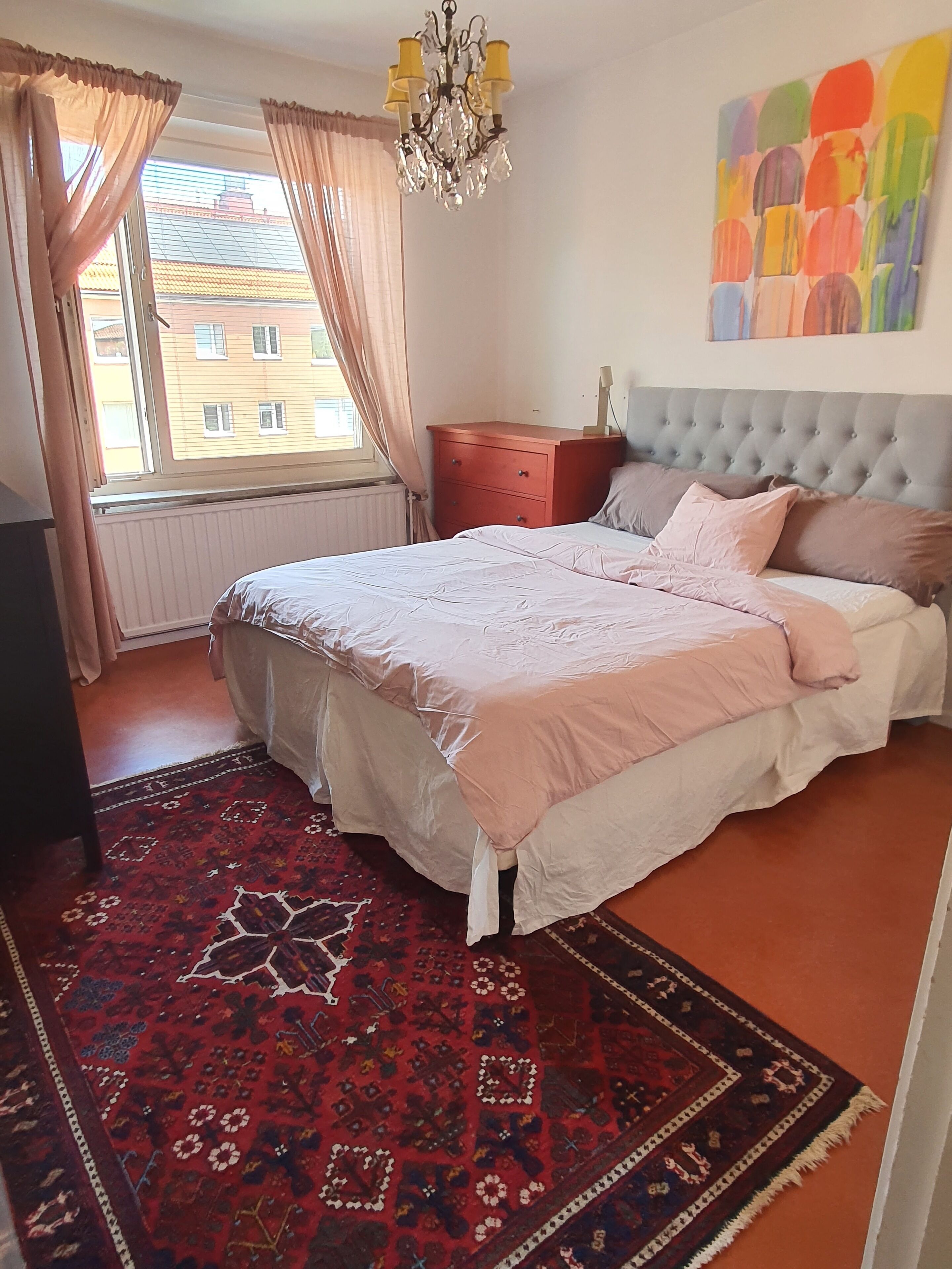 Ein Schlafzimmer mit Bett, Nachttisch, Schminktisch, einer Chandelüster, einem Fenster mit Vorhängen und einem farbenfrohen, abstrakten Gemälde an der Wand.