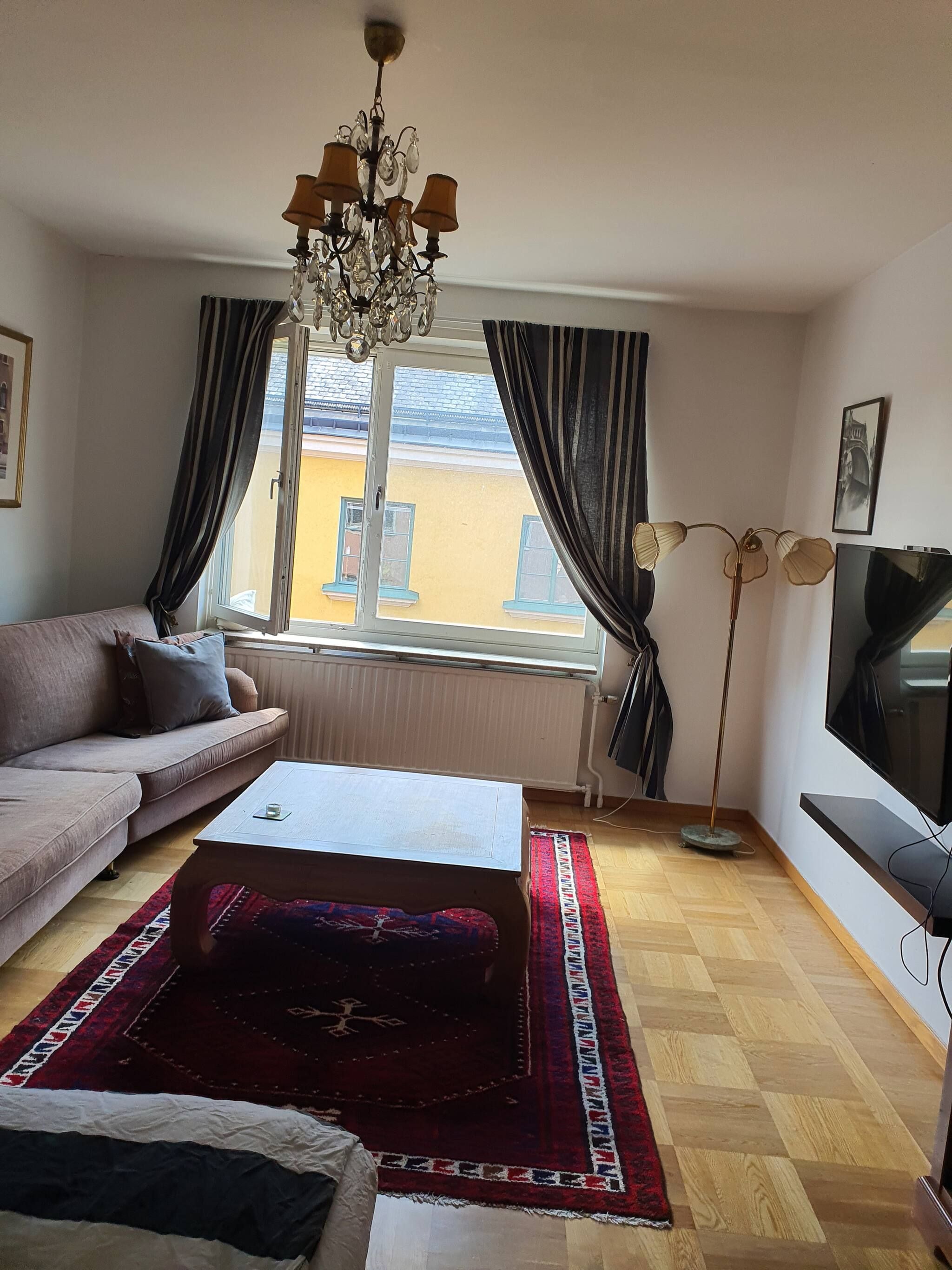 Un salón con sofá, una mesa de centro, televisor, candelabro y vistas a edificios.