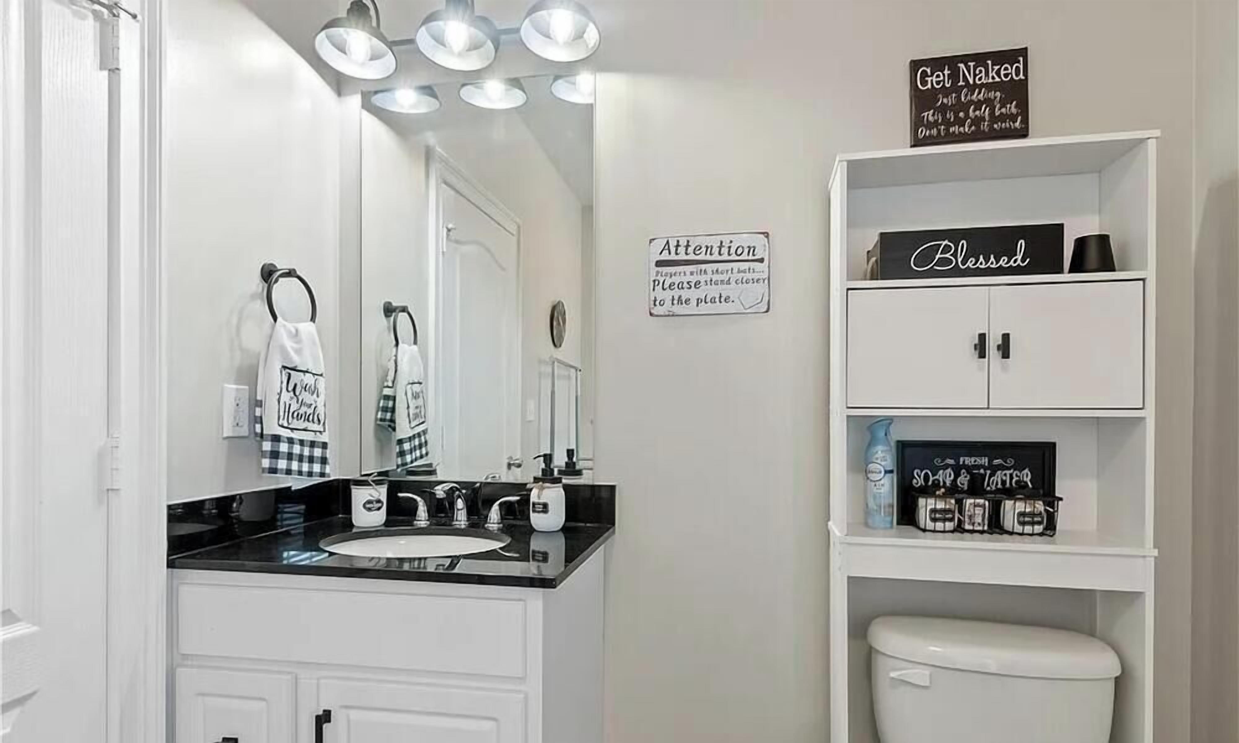 A bathroom with a white vanity, a sink, and a mirror. There are towels hanging on hooks, a soap dispenser, and a sign on the wall.