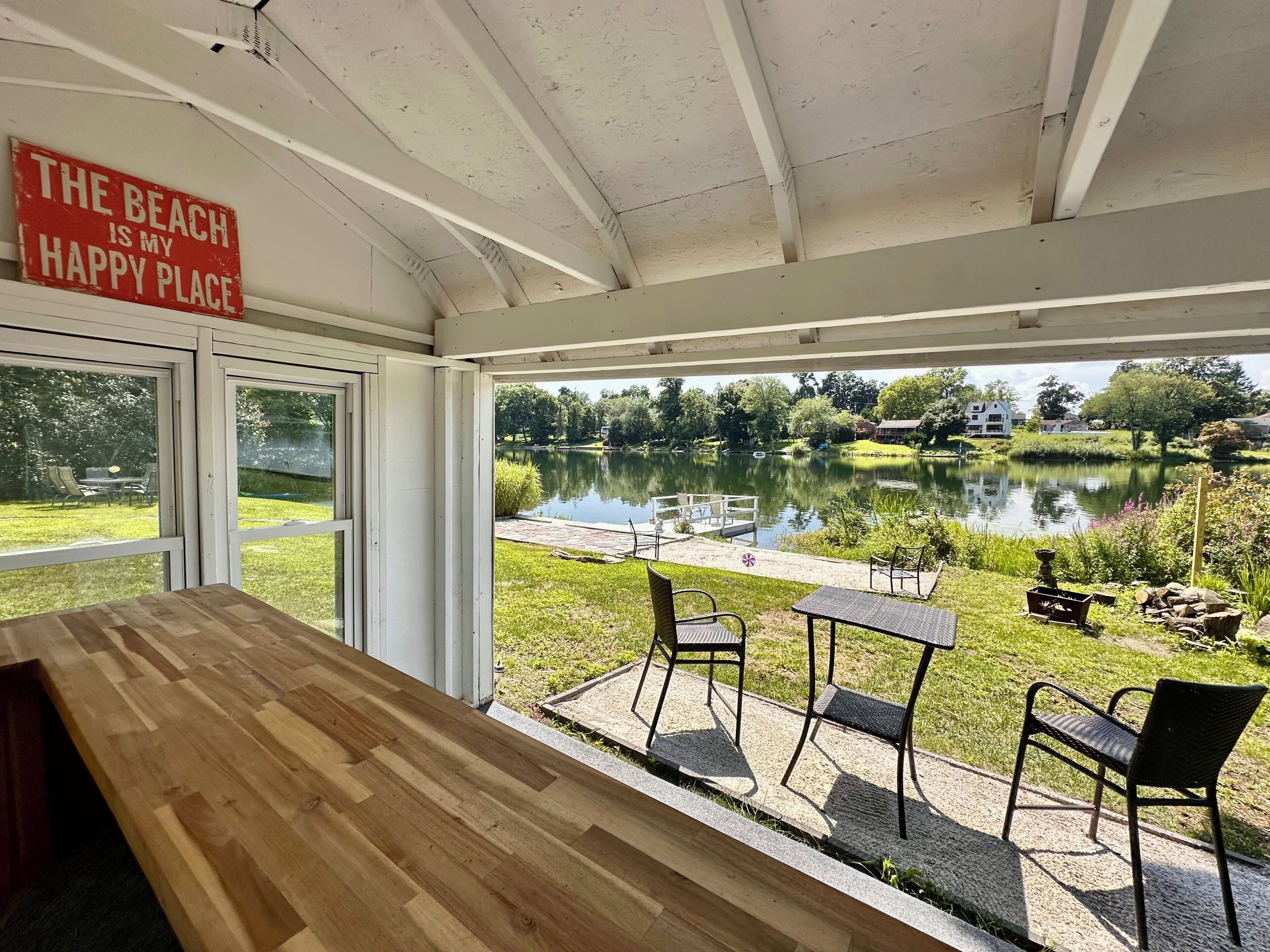 A room with a wooden table and chairs, a sign reading 'The Beach is my Happy Place,' and a view of a lake with a dock and chairs outside.