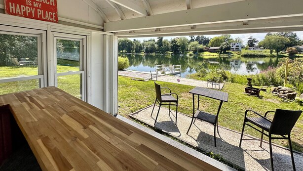 A room with a wooden table and chairs, a sign reading 'The Beach is my Happy Place,' and a view of a lake with a dock and chairs outside.