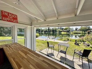 A room with a wooden table and chairs, a sign reading 'The Beach is my Happy Place,' and a view of a lake with a dock and chairs outside. - Waterfront home w/cabana, kayaks and beach! (New Fairfield)