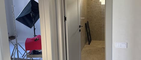 Un pasillo con una puerta que da a una habitación, una lámpara en la pared y un trípode con un atril para luces.