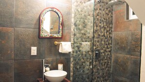 A bathroom with a stone wall, a white sink, a mirror with a colorful frame, and a glass shower enclosure. - Vrbo Property (La Estanzuela)
