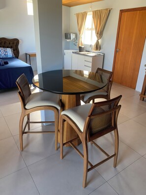 A dining area with a round table and four chairs, a bed with a blue bedspread, a kitchenette with a microwave, and a wooden door. - Pousada Santa Rosa (Imbituba)