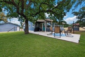 Un espace patio avec une table et des chaises, un coin salon abrité et un grand arbre qui offre de l’ombre.