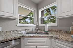 A kitchen with granite countertops, white cabinetry, a stainless steel sink, and two windows with a view of trees. - F'burg Wine Trail HQ 2BR Dog Friendly | Patio (Fredericksburg)