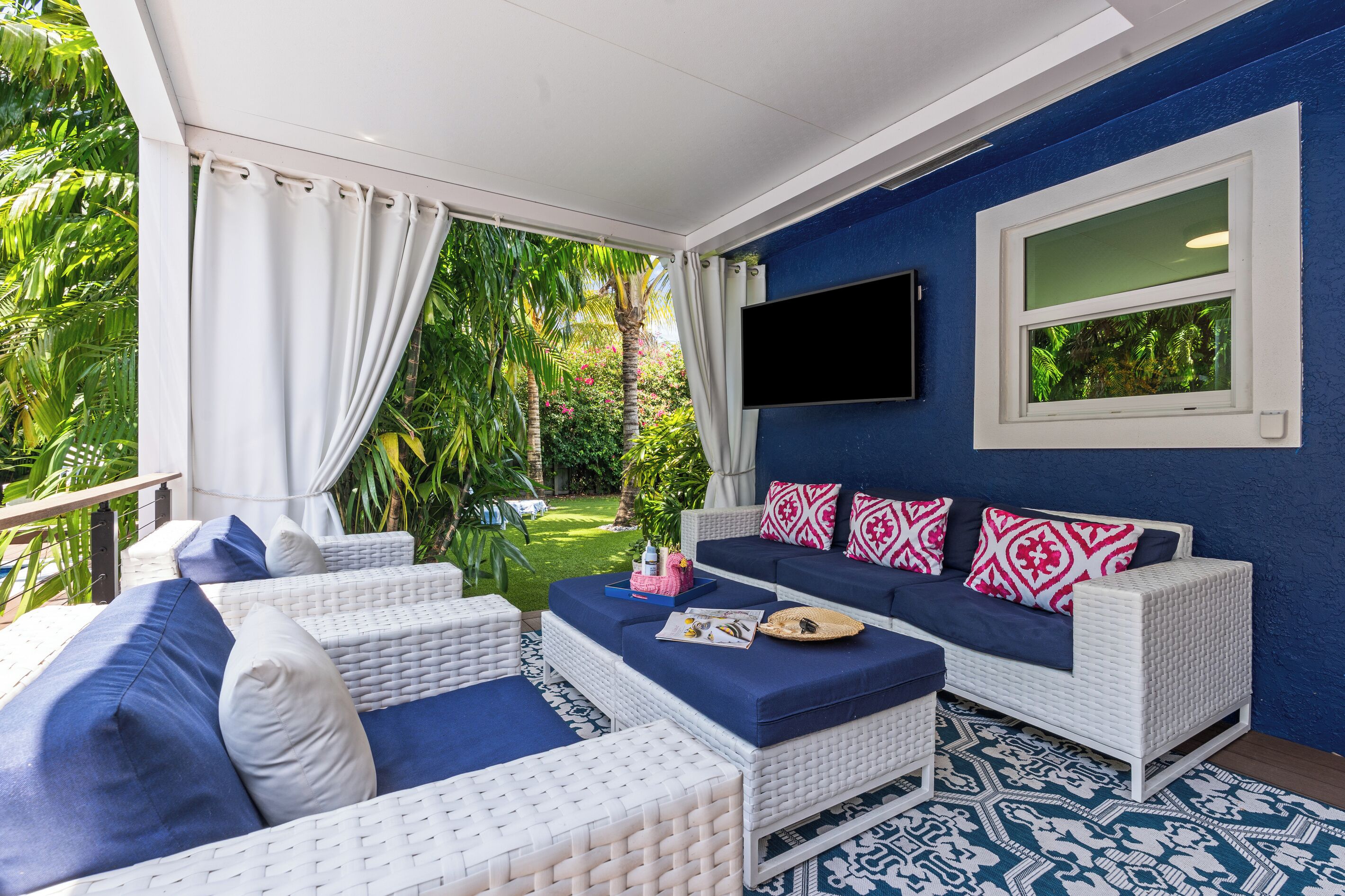 A covered outdoor seating area with white wicker furniture, a blue sofa with patterned cushions, and a large window overlooking a garden.
