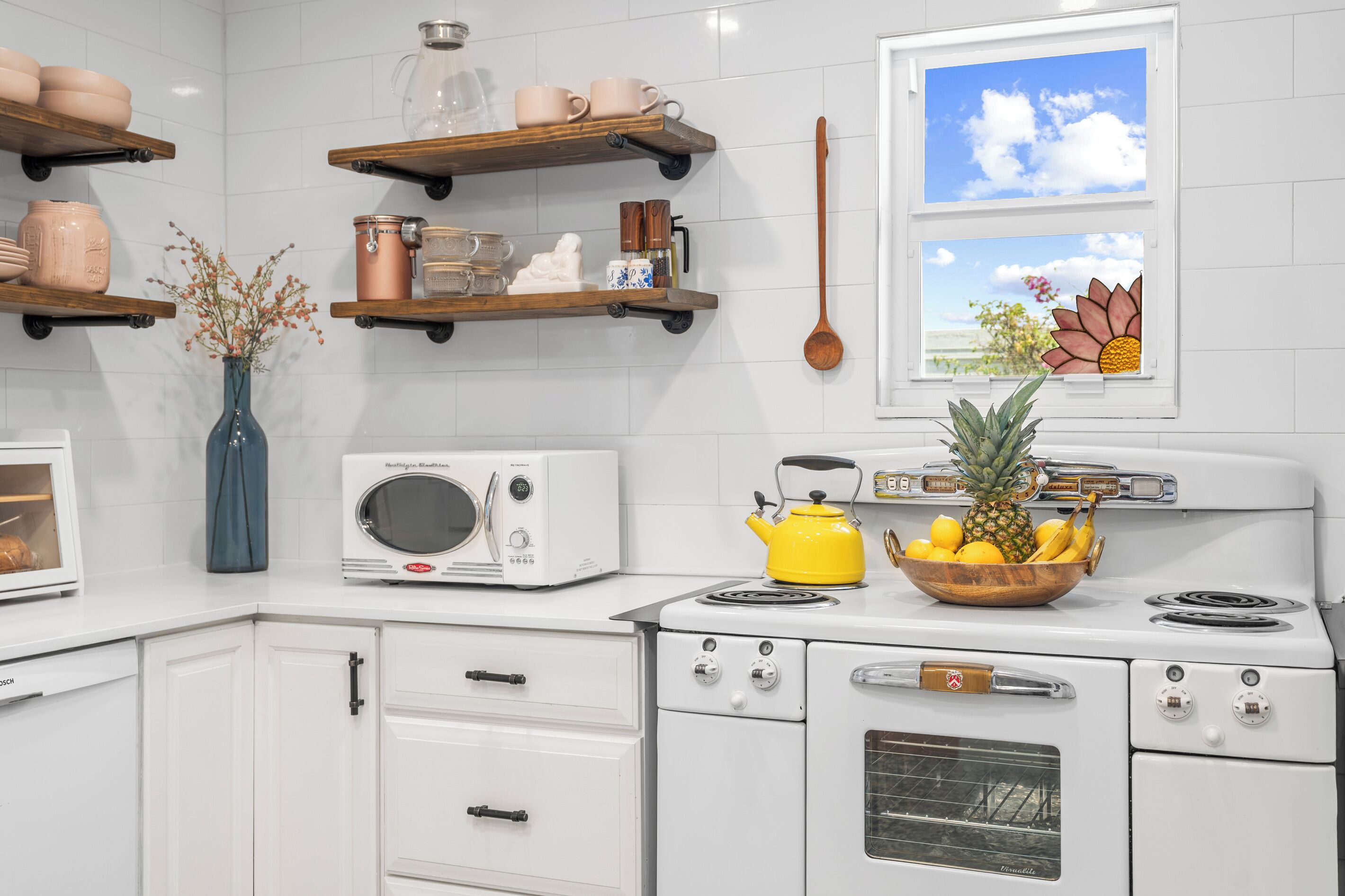 A kitchen with white cabinets, a vintage microwave, a yellow kettle, and a bowl of fruit on the stove.