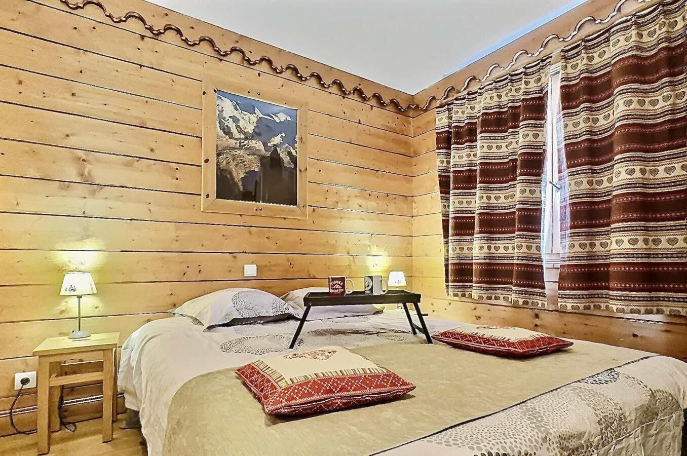 Una habitación de madera con una cama, dos mesitas de noche, una lámpara y un cuadro en la pared, además de un ventanal con cortinas estampadas.
