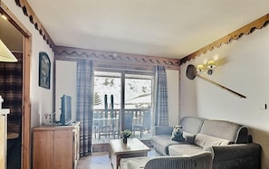 A cozy living room with a sofa, a small table, and a television. There is a window with curtains, and a door leading outside. - 380m from ski slopes, 1st floor, view mountain, balcony, tv, ski locker, 59m², Tignes (Tignes)
