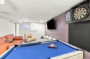 A room with a pool table, a dartboard, a neon game controller sign, a television, and a sofa.